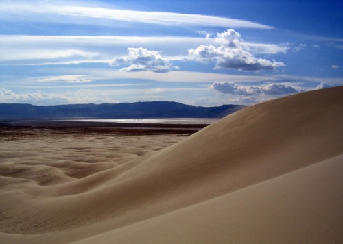 A vast desert landscape with rolling sand dunes under a blue sky with scattered clouds and distant mountains.