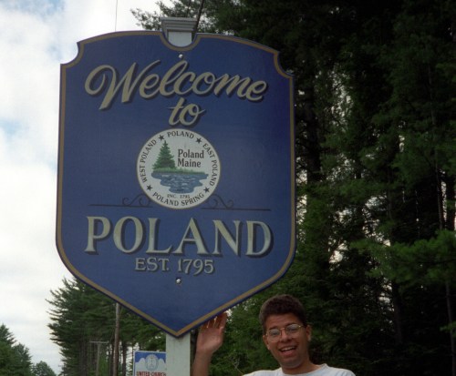 A person stands next to a blue welcome sign for Poland, Maine, established in 1795, surrounded by trees.