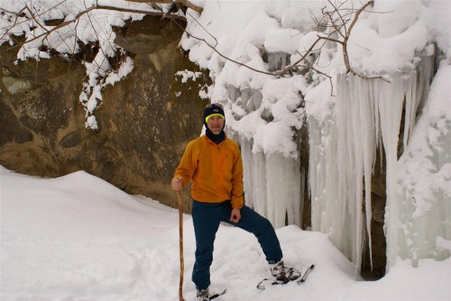 A person in winter gear stands on snow near icicles and a rocky wall, holding a walking stick.