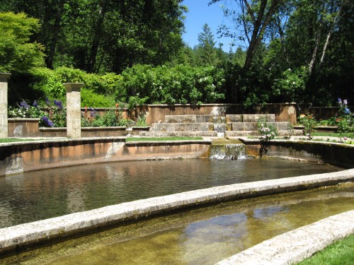 A serene garden with a stone fountain, surrounded by lush greenery and blooming flowers under a clear blue sky.