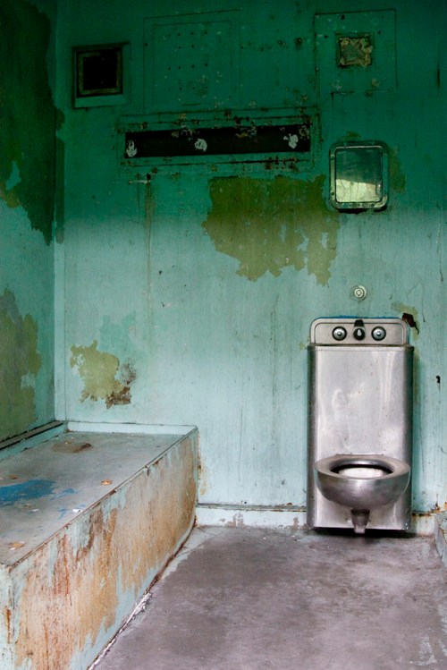 An abandoned, dilapidated bathroom with peeling turquoise paint and a metal toilet.
