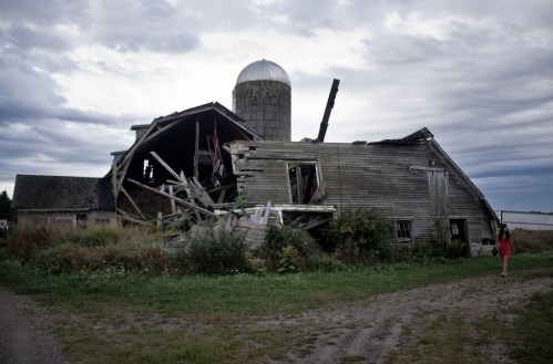 A woman walks past a dilapidated barn with a silo, surrounded by overgrown grass under a cloudy sky.