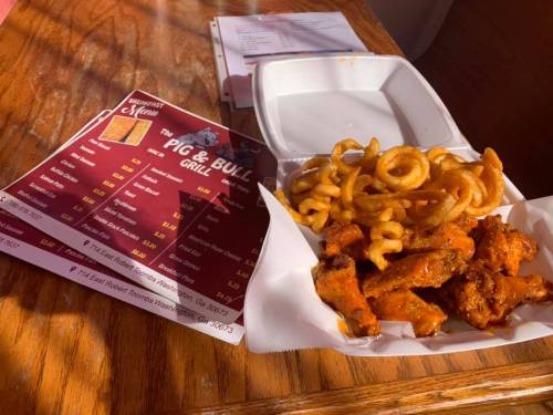 A takeout container with seasoned chicken strips and curly fries, next to a menu from The Pig & Bull Grill.