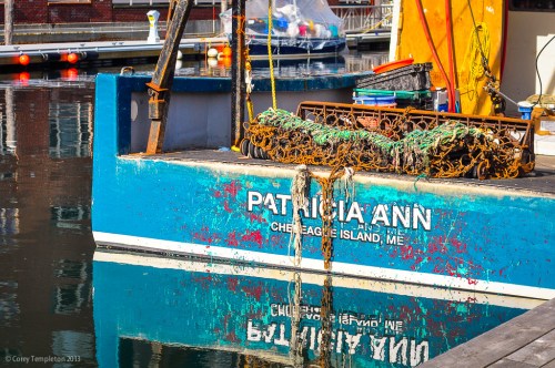 A blue fishing boat named "Patricia Ann" docked, reflecting in calm water with fishing gear on the deck.