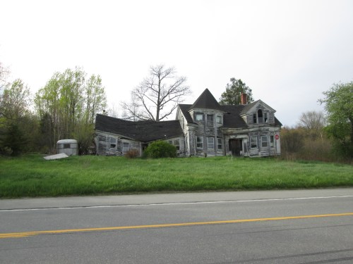 Abandoned, weathered house with a slanted roof, surrounded by overgrown grass and trees, beside a road.