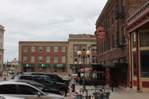 View of Lincoln Station with nearby buildings, parked cars, and outdoor seating in a city setting. Overcast sky.