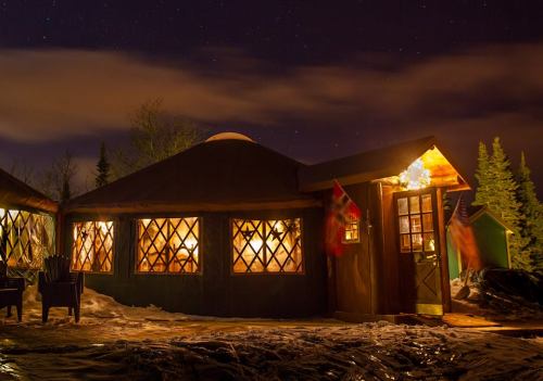 A cozy yurt illuminated at night, surrounded by snow and trees, under a starry sky. Warm light glows from the windows.