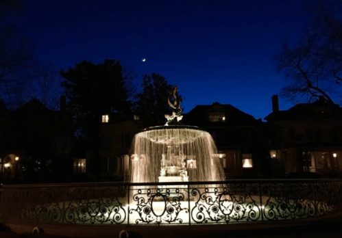 A illuminated fountain at night, surrounded by trees and historic buildings, with a crescent moon in the sky.
