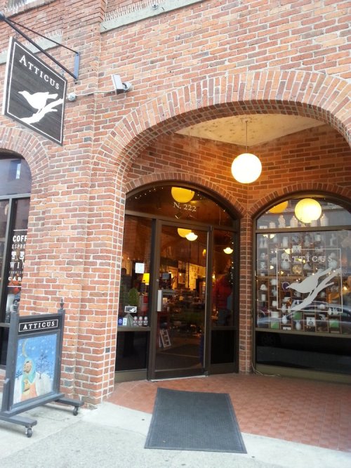 Entrance of Atticus, a brick storefront with large windows and a sign, showcasing a cozy atmosphere.