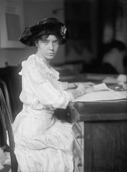 A woman in a striped dress and large hat sits at a table, writing on paper with a focused expression.
