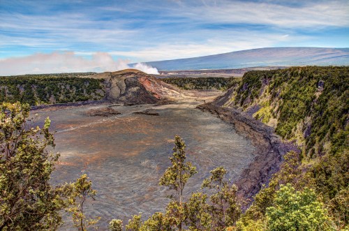 A panoramic view of a volcanic crater with smoke rising, surrounded by lush greenery and distant mountains.