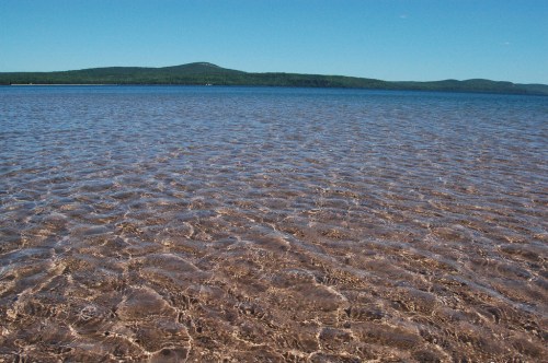 Clear water ripples over a sandy bottom, with green hills and a blue sky in the background.