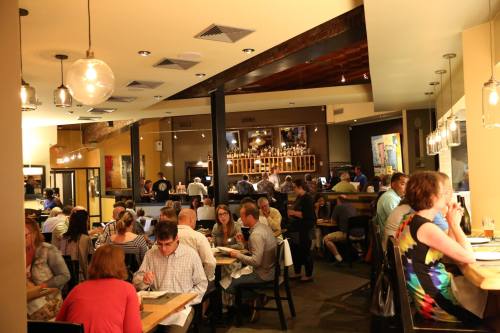 A bustling restaurant interior with diners at tables, warm lighting, and a visible kitchen in the background.