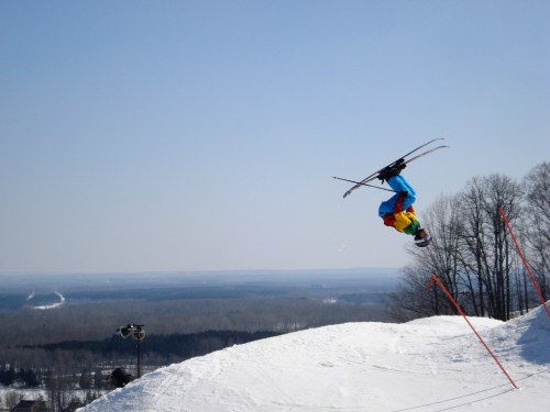 A skier performs a backflip in mid-air over a snowy slope, with trees and a distant landscape in the background.