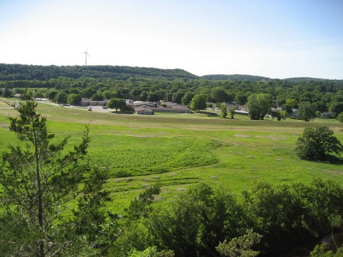 A scenic view of a green landscape with trees, a field, and a wind turbine in the distance under a clear blue sky.