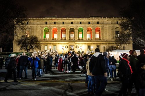 A festive crowd gathers outside a beautifully lit building decorated for the holidays, with trees and wreaths.