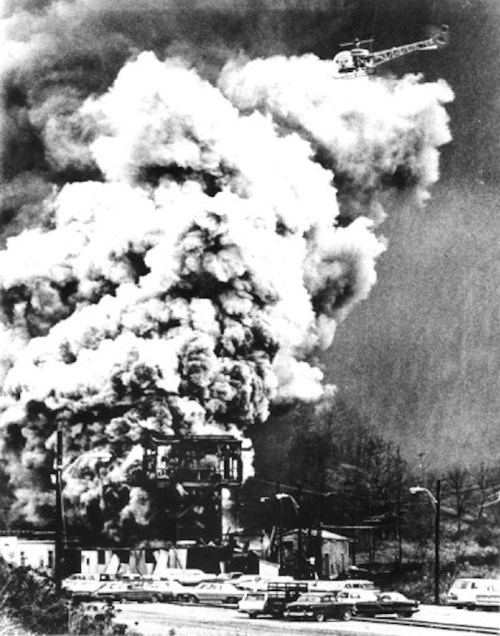 A large plume of smoke rises from a burning building, with a helicopter hovering above and cars parked nearby.