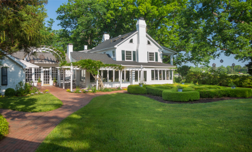 A charming white house with a landscaped garden and brick pathway, surrounded by lush green trees.