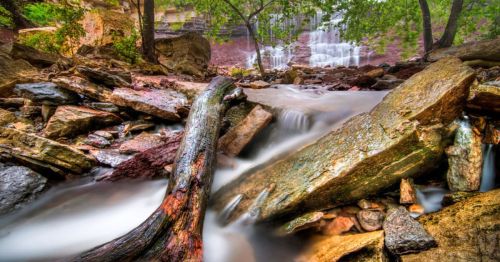 A serene waterfall cascades over rocks, surrounded by lush greenery and a flowing stream in a tranquil natural setting.