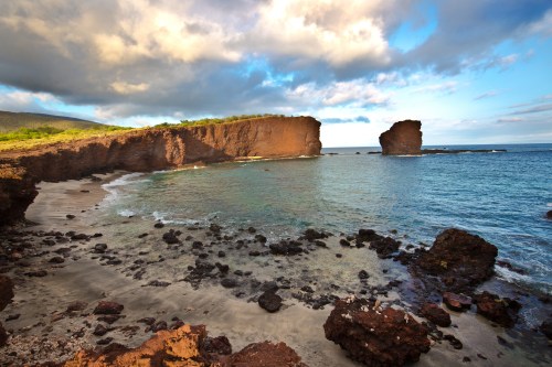 Scenic coastal view with rocky cliffs, sandy beach, and calm blue waters under a partly cloudy sky.