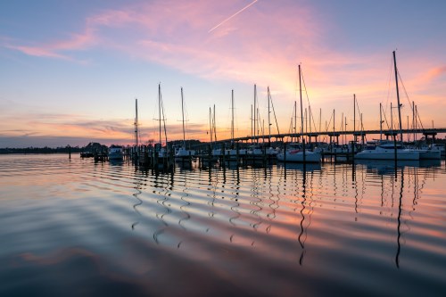 A serene sunset over a marina, with sailboats reflecting on calm waters and a colorful sky.