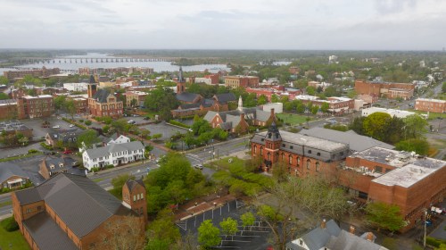Aerial view of a small town with historic buildings, green trees, and a river in the background under a cloudy sky.