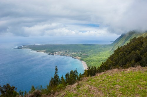 A scenic coastal view with lush green hills, a sandy beach, and cloudy skies over the ocean.