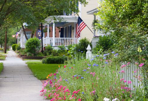 A charming neighborhood with colorful flowers lining the sidewalk and American flags adorning the porches of houses.