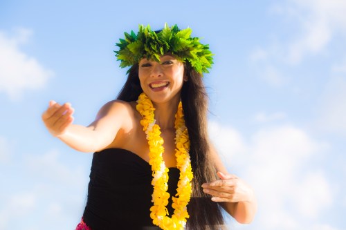 A woman in a black dress and flower lei dances joyfully outdoors, wearing a leafy crown against a blue sky.