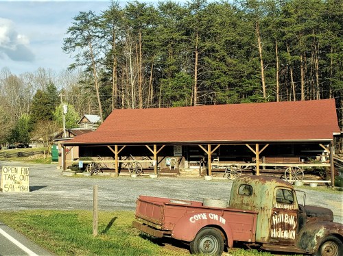 A rustic building with a red roof, surrounded by trees, featuring a vintage truck and a sign for takeout.