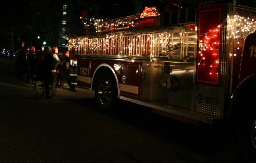 A decorated fire truck with lights parked on a street, with people in festive attire nearby.