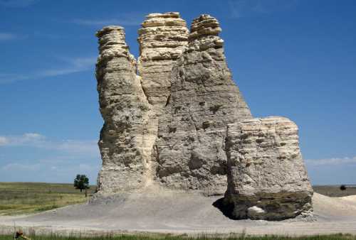 A tall, unique rock formation with layered, weathered cliffs against a clear blue sky and grassy landscape.