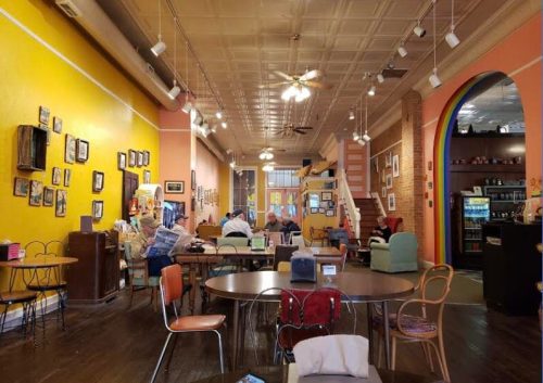 Cozy café interior with colorful walls, tables, and chairs, featuring artwork and patrons enjoying the space.