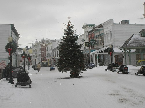 A snowy street scene featuring a Christmas tree and decorated buildings, with people and snowmobiles in the foreground.