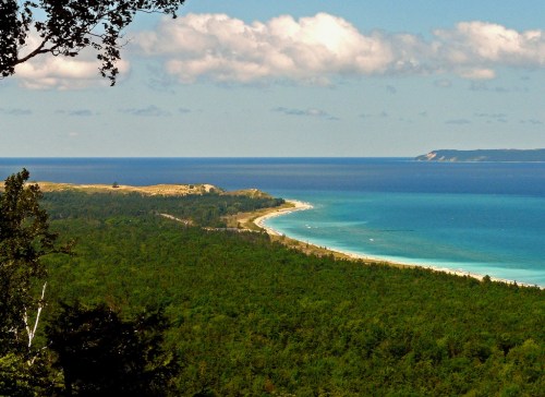 A scenic view of a coastline with lush greenery, blue waters, and distant hills under a partly cloudy sky.