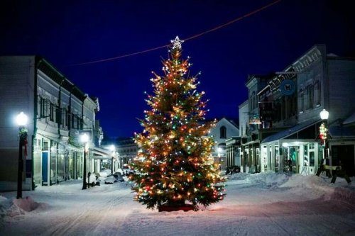 A brightly lit Christmas tree stands in a snowy street, surrounded by charming buildings at night.