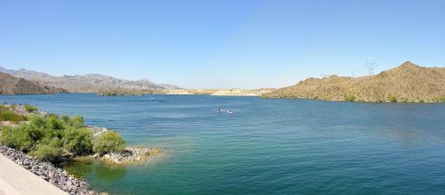 A serene landscape featuring a calm blue lake surrounded by mountains and greenery under a clear blue sky.