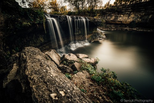 A serene waterfall cascades over rocky cliffs into a calm pool, surrounded by lush greenery and autumn foliage.
