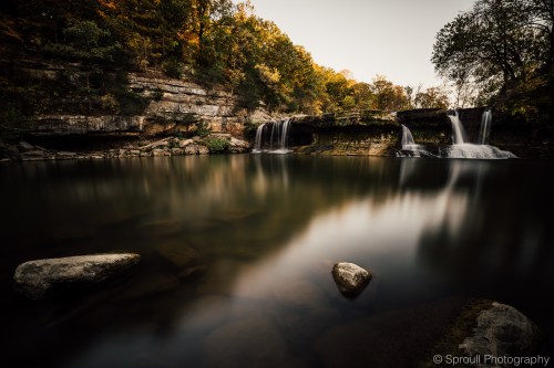 A serene waterfall cascades into a calm pool, surrounded by lush greenery and rocky terrain under a golden sky.
