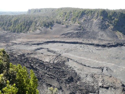 A vast, barren landscape of hardened lava flows surrounded by green vegetation and steep cliffs in the distance.