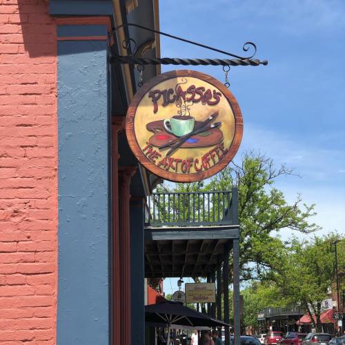 Sign for "Picasso's: The Art of Coffee" hanging from a building, with a blue sky and trees in the background.
