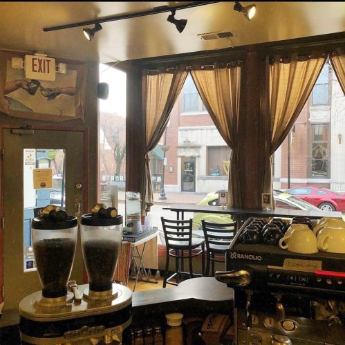 A cozy café interior with large windows, curtains, and coffee equipment, overlooking a snowy street outside.