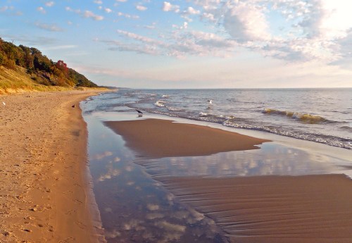 A serene beach scene with gentle waves, soft sand, and a cloudy sky reflecting on the water.