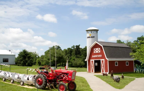 A red barn and tractor in a sunny farmyard, with a silo and playful goats nearby.