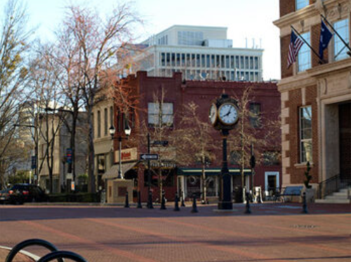 A street view featuring a vintage clock, brick buildings, and trees in a downtown area on a clear day.