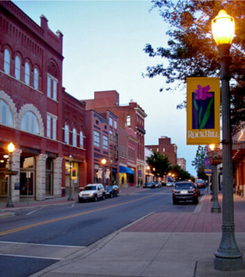 A quiet street in Rock Hill, featuring brick buildings, street lamps, and banners, during twilight.