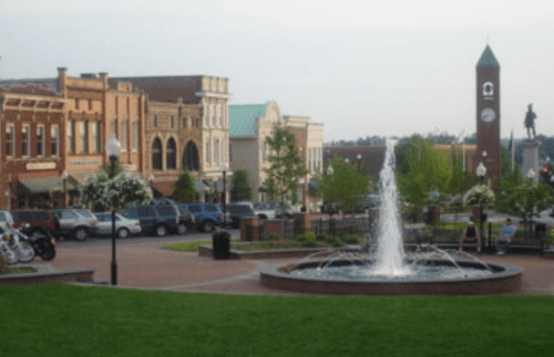 A town square featuring a fountain, historic buildings, trees, and parked cars under a clear sky.
