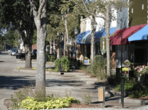 A quiet street lined with trees and shops featuring colorful awnings on a sunny day.