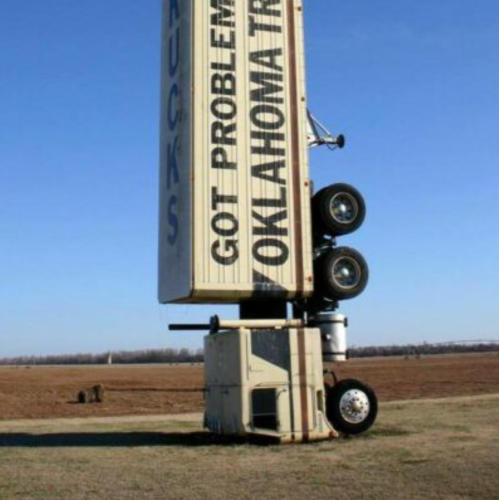 A large truck trailer is upright on its end, with wheels at the top, in a flat, open field under a clear blue sky.