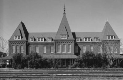 Historic brick building with multiple peaked roofs and large windows, surrounded by greenery and railway tracks.